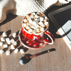 A cup of cocoa with marshmelow on an grey wooden background with a spoon and ebook, frame format.