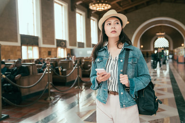 Smiling asian woman traveler standing carrying backpack in union station hall. Pretty young lady wearing straw hat and holding smartphone checking train online timetable. Tourism and travel concept