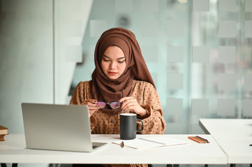 Islam woman working with laptop and holding a coffee mug and glasses.