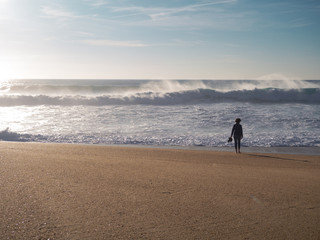 Naklejka premium A young woman walking along the wide beach with dangerous curving waves; stormy sea in clear weather Nazare; leisure activity; boots in hand; crystal clear drops of seawater fly in the air; Portugal