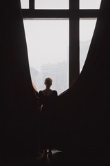 A beautiful slender young woman with light short hair in a black dress stands at the window in a room with dark curtains. Vertical picture.
