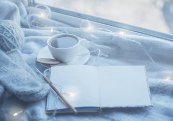 Cozy winter still life: mug of hot tea and book with warm plaid on windowsill against snow landscape from outside.