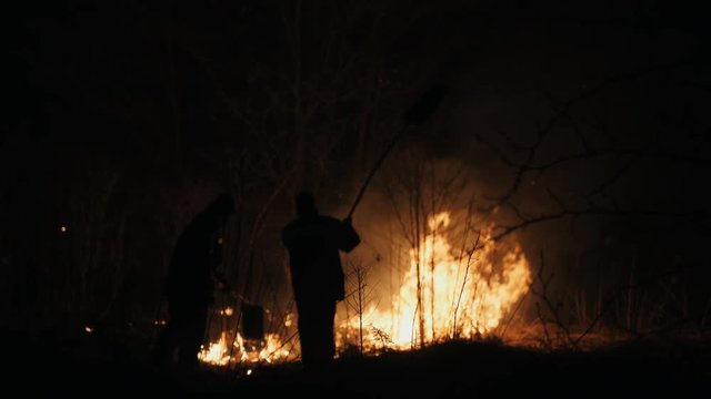 Two firefighters with fire flappers extinguish a fire at night