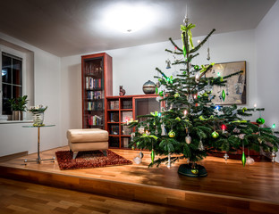Richly decorated Christmas tree in a cool, modern home library with wooden floor and wooden furniture.