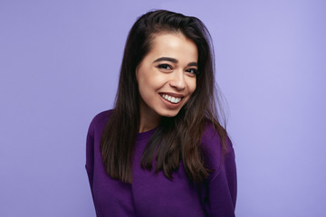 Fototapeta premium Portrait of pretty adult girl, broad smile, wears purple sweatshirt, tilts head and looks with joy, isolated over violet wall