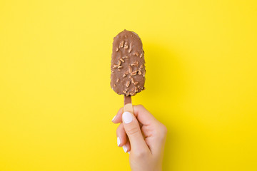 Young woman hand holding ice cream with nuts and chocolate glaze on bright yellow background. Empty...
