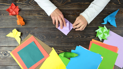 Man making pink paper crane. Multicolored paper sheets for artwork, top view. How to make origami orizuru instructions.