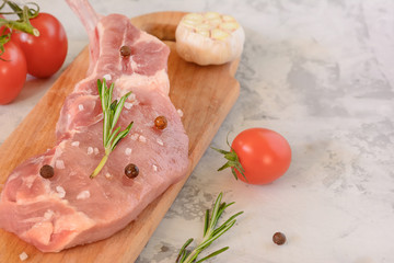 A piece of fresh pork meat lies on a cutting board, alongside garlic and tomatoes. Preparation for cooking steak. Copy space on light gray background.