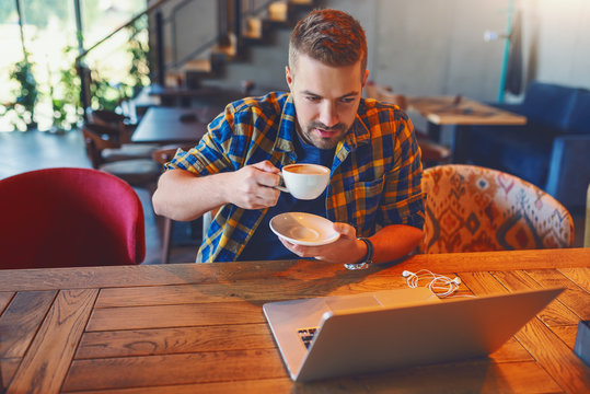 Young Caucasian blogger in plaid shirt drinking coffee and looking at laptop while sitting in coffee shop.