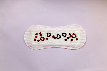 Period pad with red heart shaped sequins symbolizing menstruation. Top view, lilac background.