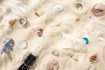 top view of seashells, bottle caps, scattered cigarette butts, plastic cups, glass bottle and candy wrapper on sand