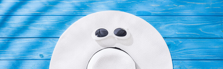 panoramic shot of white floppy hat with black ribbon and sunglasses on blue wooden background