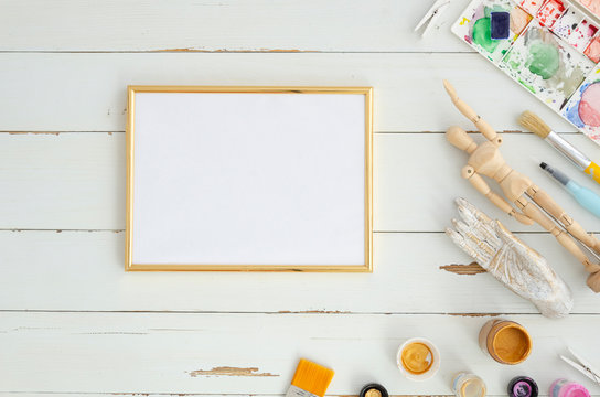 Creative artist workplace mockup. Top view on table with blank sheet of paperin golden frame with paintbrushes. Copy space for text on white wooden planks background