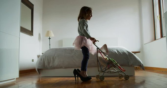 Authentic shot of little girl is playing with stroller toy and doll wearing her mother high heeled shoes in parents bedroom.