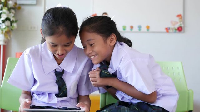 Two Thai cute elementary school girls, long hair in a white shirt dress, skirt and green plaid tie are playing games on the tablet easily in school.