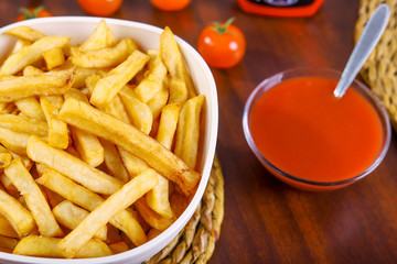 French fries in white bowl on a braided pad on wooden table with tomato and ketchup next to it