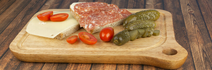 A slice of bread with salami and a slice of bread with cheese, while still small cocktail tomatoes and pickled cucumbers, served on a wooden board