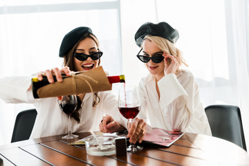 brunette and blonde happy women in black berets and sunglasses pouring red wine and reading magazine