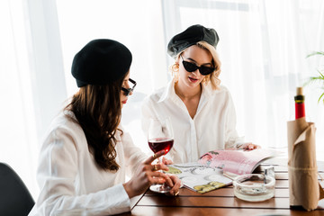 brunette and blonde women in black berets and sunglasses drinking red wine and reading magazine