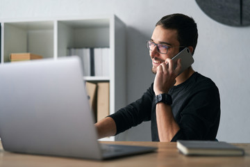 Young man working with phone and computer, receiving phone call, talking with partners 