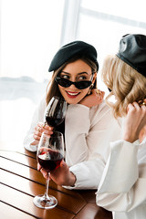 elegant smiling brunette woman in black beret and sunglasses drinking red wine at wooden table with blonde friend