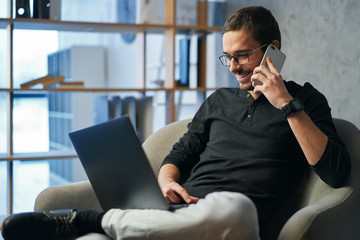 Young man working with phone and computer, receiving phone call, talking with partners 