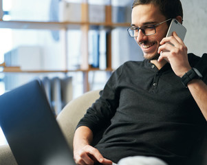 Young man working with phone and computer, receiving phone call, talking with partners 