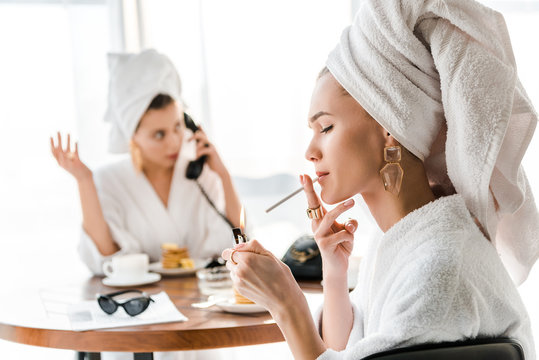 selective focus of stylish woman in bathrobe and jewelry with towel on head lighting up cigarette while friend talking on phone