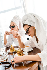 Obraz na płótnie Canvas stylish women in bathrobes, sunglasses and jewelry with towels on heads eating pancakes for breakfast