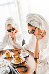 Stylish smiling woman in bathrobe, sunglasses and jewelry with towel on head smoking cigarette and reading newspaper with friend at morning