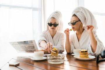 Stylish women in bathrobes, sunglasses and jewelry with towels on heads smoking cigarette and reading newspaper while eating pancakes
