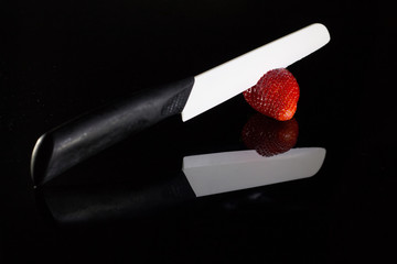 A white blade knife cuts a fresh strawberry and its reflection with black background.