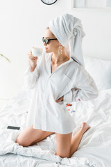 young stylish woman in shirt, sunglasses, jewelry and with towel on head sitting on bed while drinking coffee