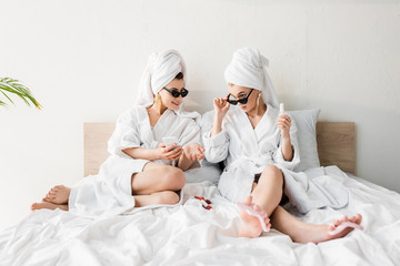 stylish women in bathrobes and sunglasses, towels and jewelry lying in bed, doing pedicure and using smartphone