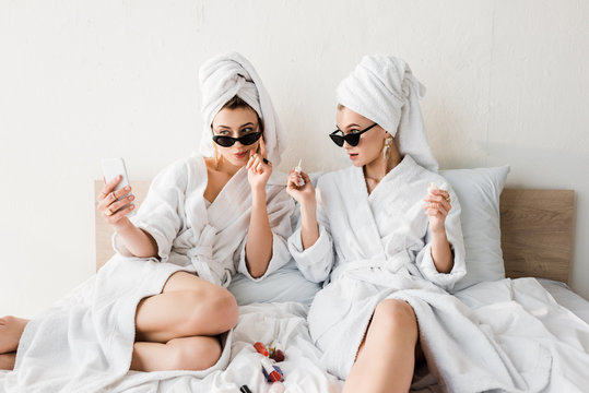 stylish women in bathrobes and sunglasses, towels and jewelry doing pedicure and taking selfie in bed