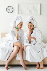 stylish smiling girls in earrings, bathrobes and with towels on heads talking while sitting on bed