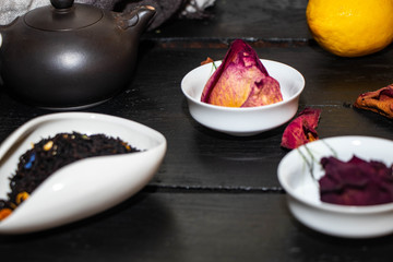Special white bowl for dry, crumbly tea. Next to lemon and cups for Chinese tea. in cups dry roses and rose petals on a black background. Brown, Chinese teapot. 