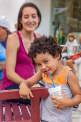 A small boy with curly hair smirks at the camera. A toddler stands at the edge of the bench looking at the camera while mommy smiles in the background setting downtown.