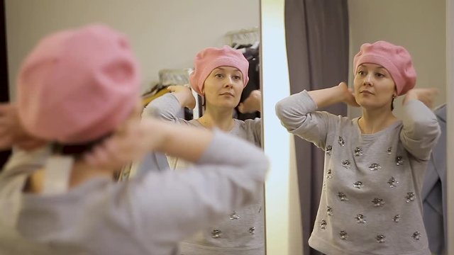 Young woman trying on clothes. Attractive caucasian woman looks in the mirror, turning her head, trying on a hat in a fitting room of a clothing store.
