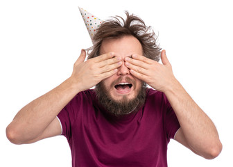 Crazy bearded Man with funny Haircut in birthday cap, isolated on white background. Happy Guy covers his eyes with his hands - can not see. Holidays concept.