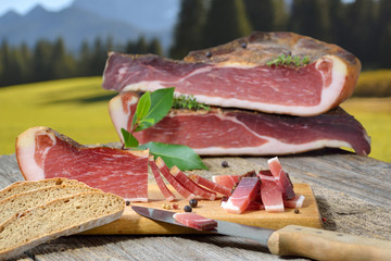 Südtiroler Speckjause mit Vinschgerl auf einem Holztisch vor Bergen in den Alpen – Typical South Tyrolean bacon snack with local rye bread lying on a rustic table in front of mountains of the alps