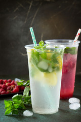Refreshing summer drink lemon with mint, ice and raspberry with basil, ice. Glasses with cold and healthy beverage on a dark stone background. Copy space.