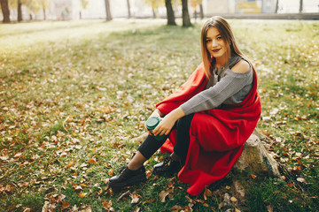 pretty cute girl with make up, dressed in gray sweater sitting in a autumn park on a red plaid and drinking a coffee