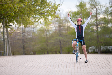 Fototapeta na wymiar Young man in helmet and sportswear raising hands and celebrating victory while riding bicycle on pavement in park