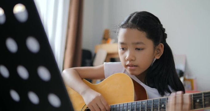 Asian little girl learning to play basic guitar by using acoustic guitar for beginner music instrumental self studying at home