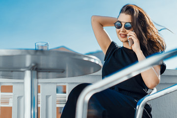 Stylish young woman sitting outdoors on balcony and talking by mobile phone.