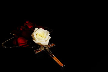 Crucifix inlaid with wood (souvenir) with gold chain, white rose and red rose petals on black background. Symbolic concept — faith, Christianity, resurrection, life. Play of light and shadow. - Powered by Adobe