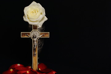 Tin cup with white rose, crucifix inlaid with wood (souvenir) and red rose petals on black background. Symbolic concept — faith, Christianity, resurrection, life. Minimal style.