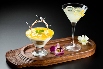 Composition of dessert and cocktail with lemon on a wooden plate on a dark background.