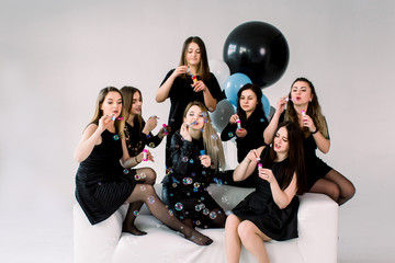 Happy beautiful women celebrating birthday or hen party, sitting on white sofa indoor and blowing soap bubbles. Birthday party, balloons, holiday